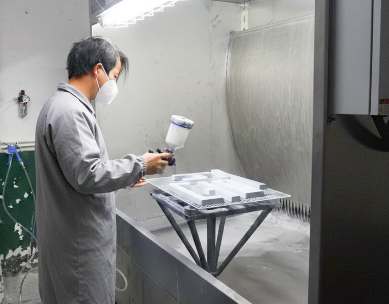 A technician wearing a protective mask uses a gravity-feed spray gun to coat industrial parts inside a water wash spray booth.