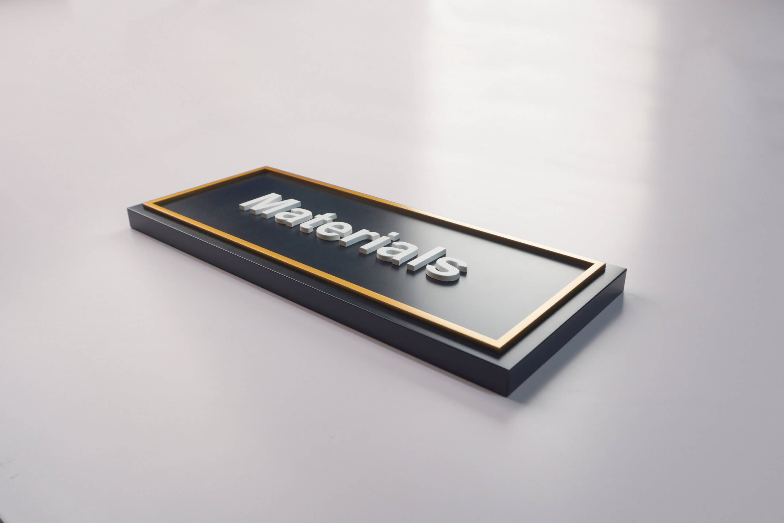 Close-up of a dark gray rectangular sign with a gold border and raised white 3D lettering that says **“Materials”**, on a clean white background, featuring a minimalist modern design style.