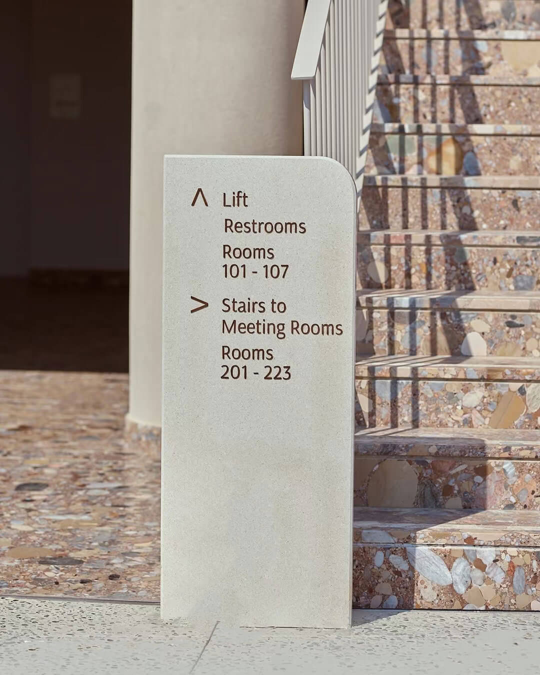A minimalist light grey stone pylon sign next to a staircase. It features an up arrow for "Lift", "Restrooms", and "Rooms 101-107", and a right arrow for "Stairs to Meeting Rooms" and "Rooms 201-223", set against colorful terrazzo stairs.
