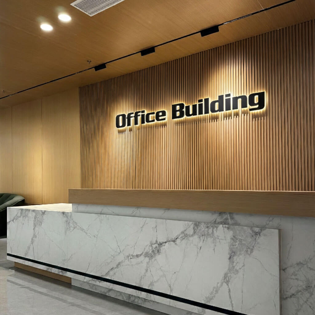 A modern office building reception area featuring a marble counter and a vertical wood slat feature wall with backlit black 3D lettering that reads "Office Building."