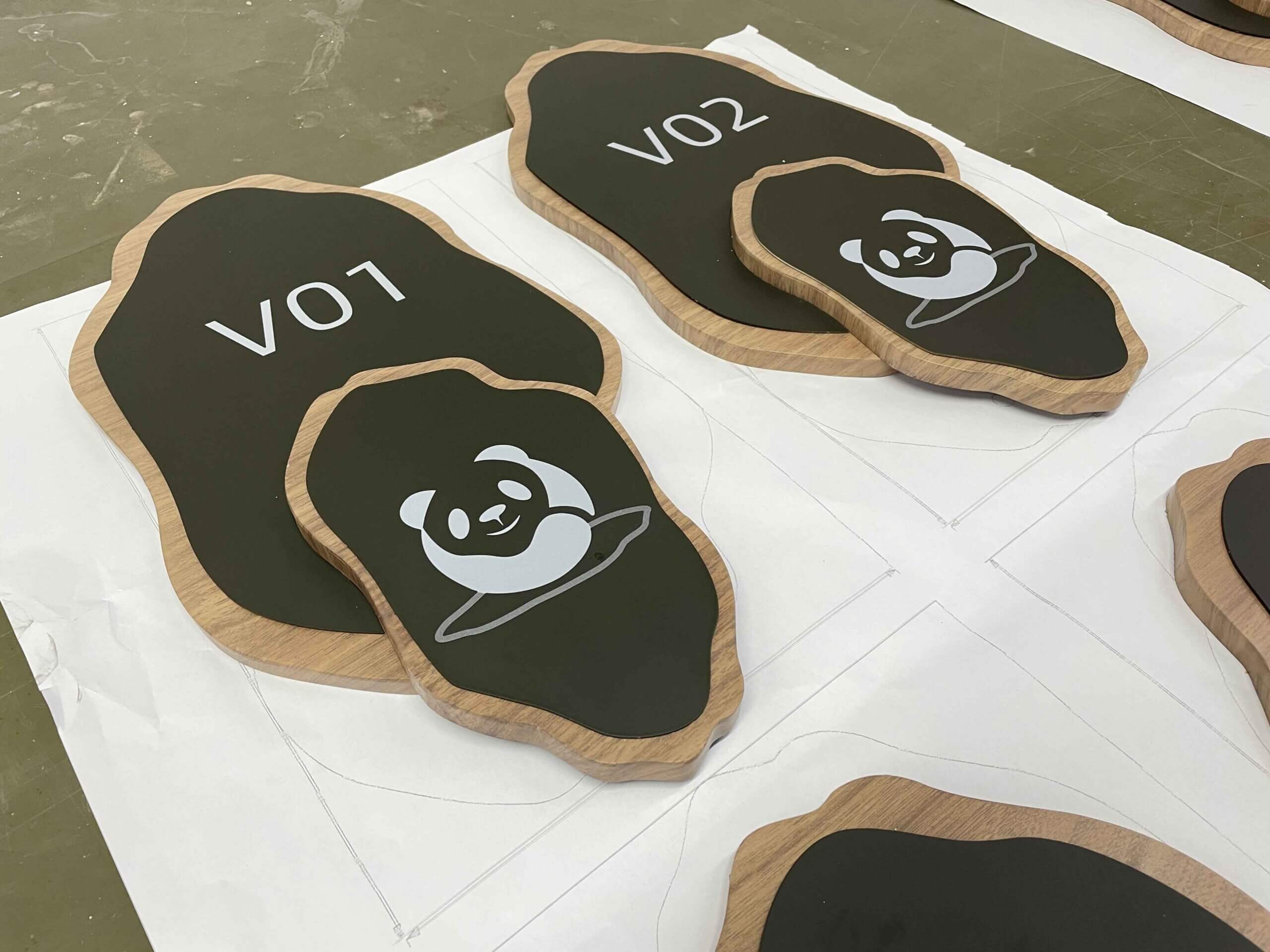 A minimalist translucent directory sign mounted on a white wall, featuring black arrows and text above a colorful 3D floor plan. A potted plant and a wooden bench are positioned next to it.