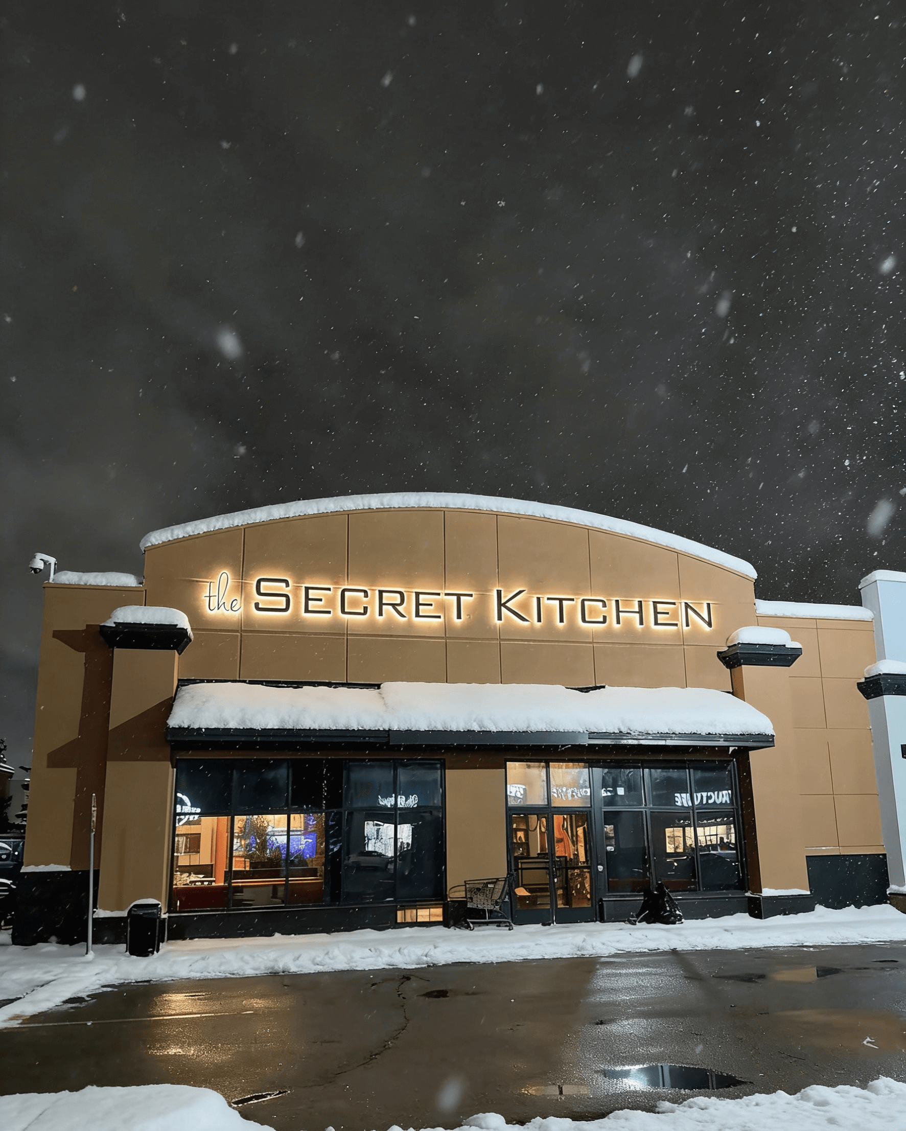 The exterior of 'The Secret Kitchen' restaurant at night in the snow, showcasing 3D metal letters with warm halo backlighting.
